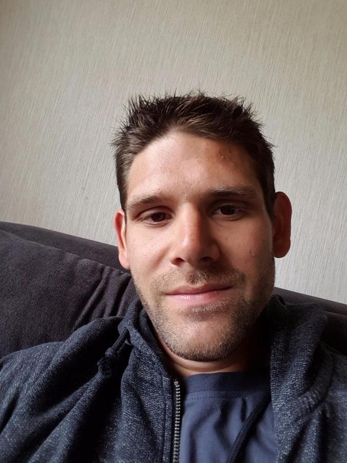 Ein Mann sitzt auf einem Sofa und lächelt in die Kamera. Er trägt einen dunklen Pullover.