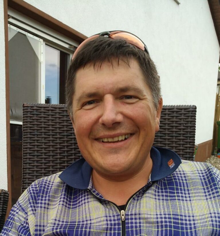 Ein lächelnder Mann mit kurz geschnittenen Haaren und einer Sonnenbrille auf dem Kopf. Er trägt ein blau-gelbes kariertes Hemd.