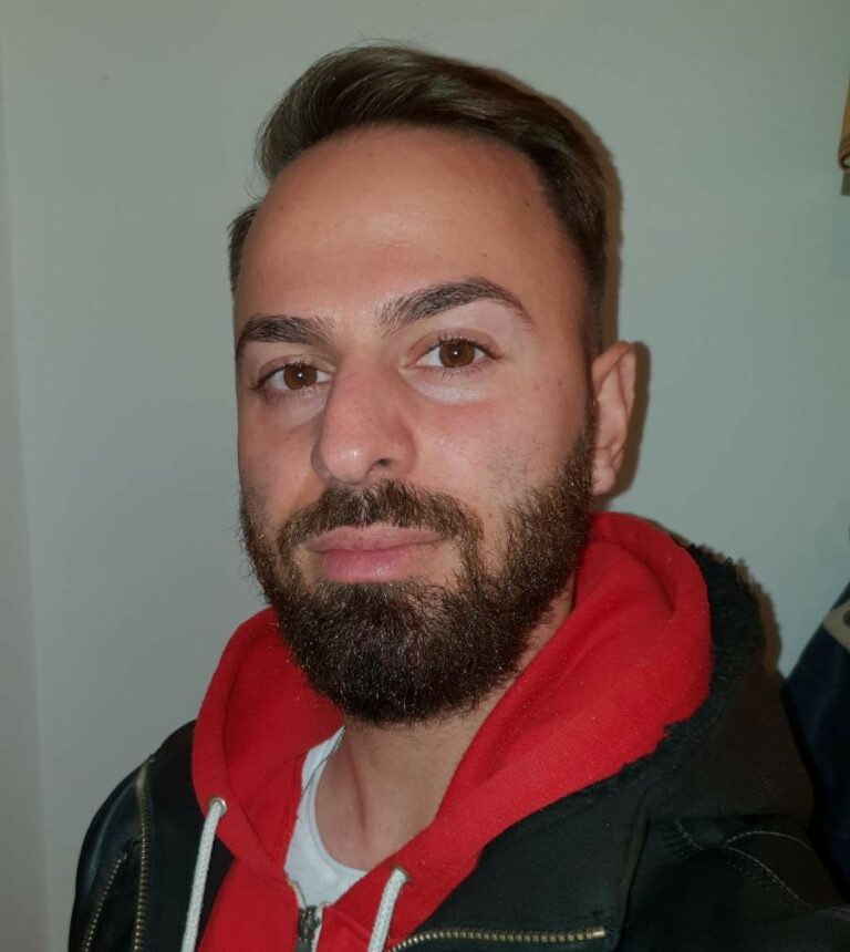 Ein Mann mit braunem Haar und Bart, trägt ein rotes Hoodie und eine schwarze Jacke.