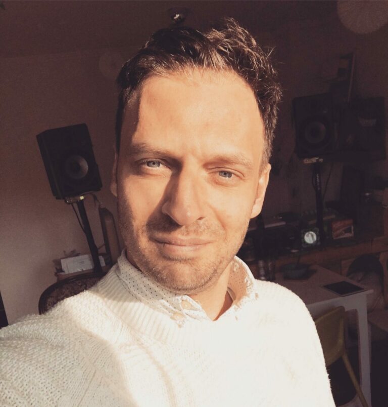 Ein Mann mit kurzem Haar und einem weißen Pullover steht in einem Raum mit Lautsprechern im Hintergrund.