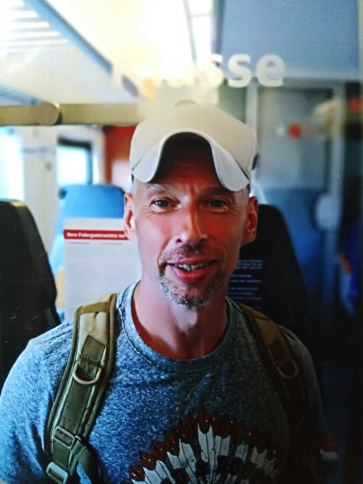 Ein Mann mit einem freundlichen Lächeln im Zug. Er trägt eine weiße Baseballkappe und einen Rucksack.