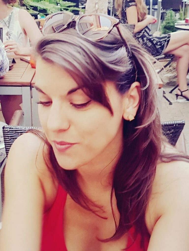 Eine junge Frau mit langen braunen Haaren sitzt im Freien. Sie trägt ein rotes Tanktop und Sonnenbrille auf dem Kopf.