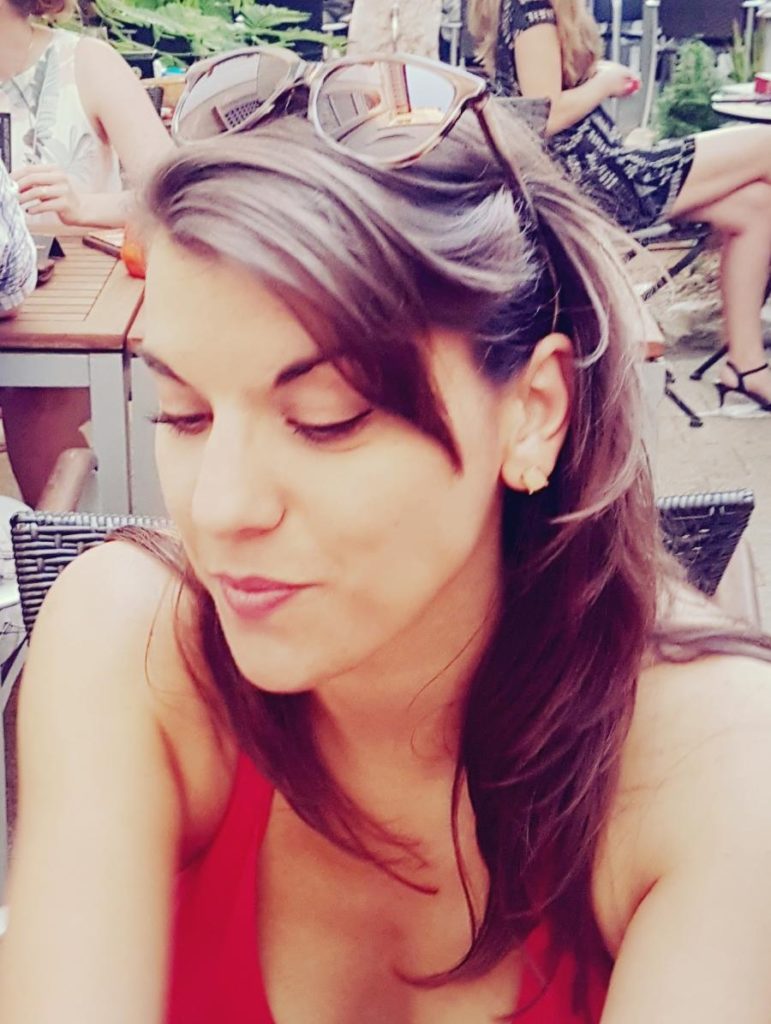 Eine junge Frau mit langen braunen Haaren sitzt im Freien. Sie trägt ein rotes Tanktop und Sonnenbrille auf dem Kopf.