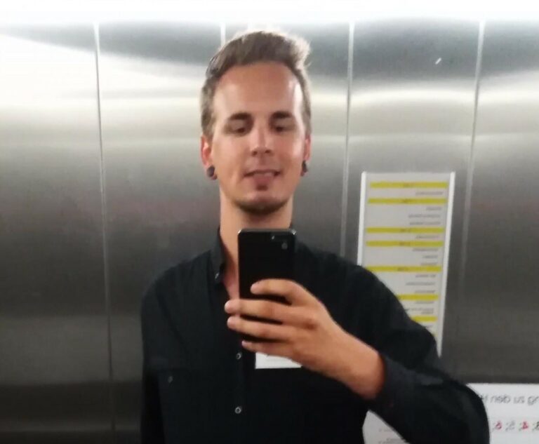 Ein Mann in einem schwarzen Hemd hält sein Telefon hoch, um ein Selfie im Aufzug zu machen. Er hat kurzes, braunes Haar und steht vor einer metallenen Wand.