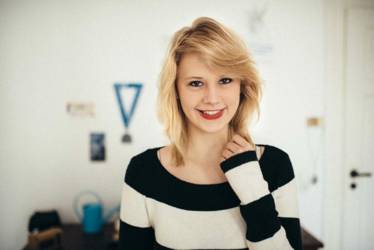 Eine junge Frau mit blondem Haar und einem schwarzen und weißen Pullover lächelt in die Kamera.