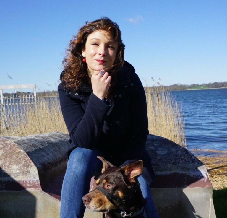 Eine Frau mit langen braunen Haaren sitzt mit einem Dobermann auf einer Bank am Wasser. Sie trägt eine schwarze Jacke und blaue Jeans.