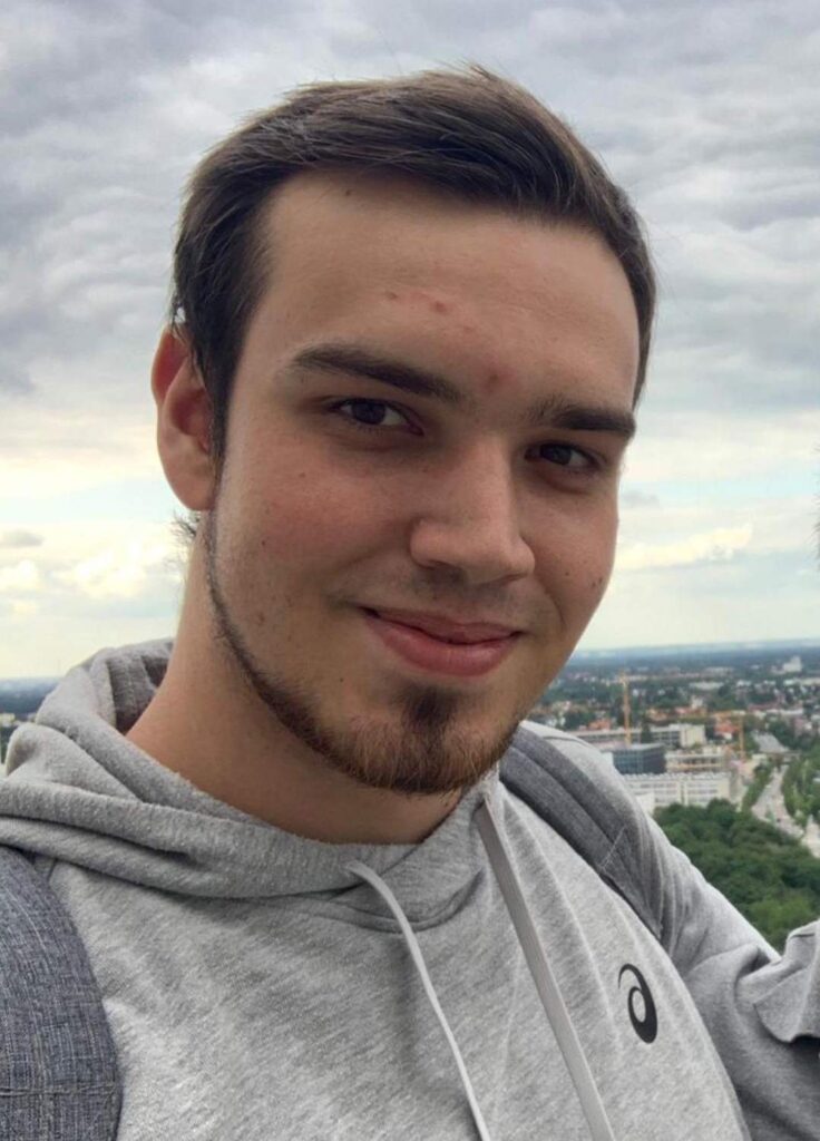 Ein junger Mann mit braunen Haaren und einem freundlichen Lächeln, im Hintergrund eine Stadtlandschaft.
