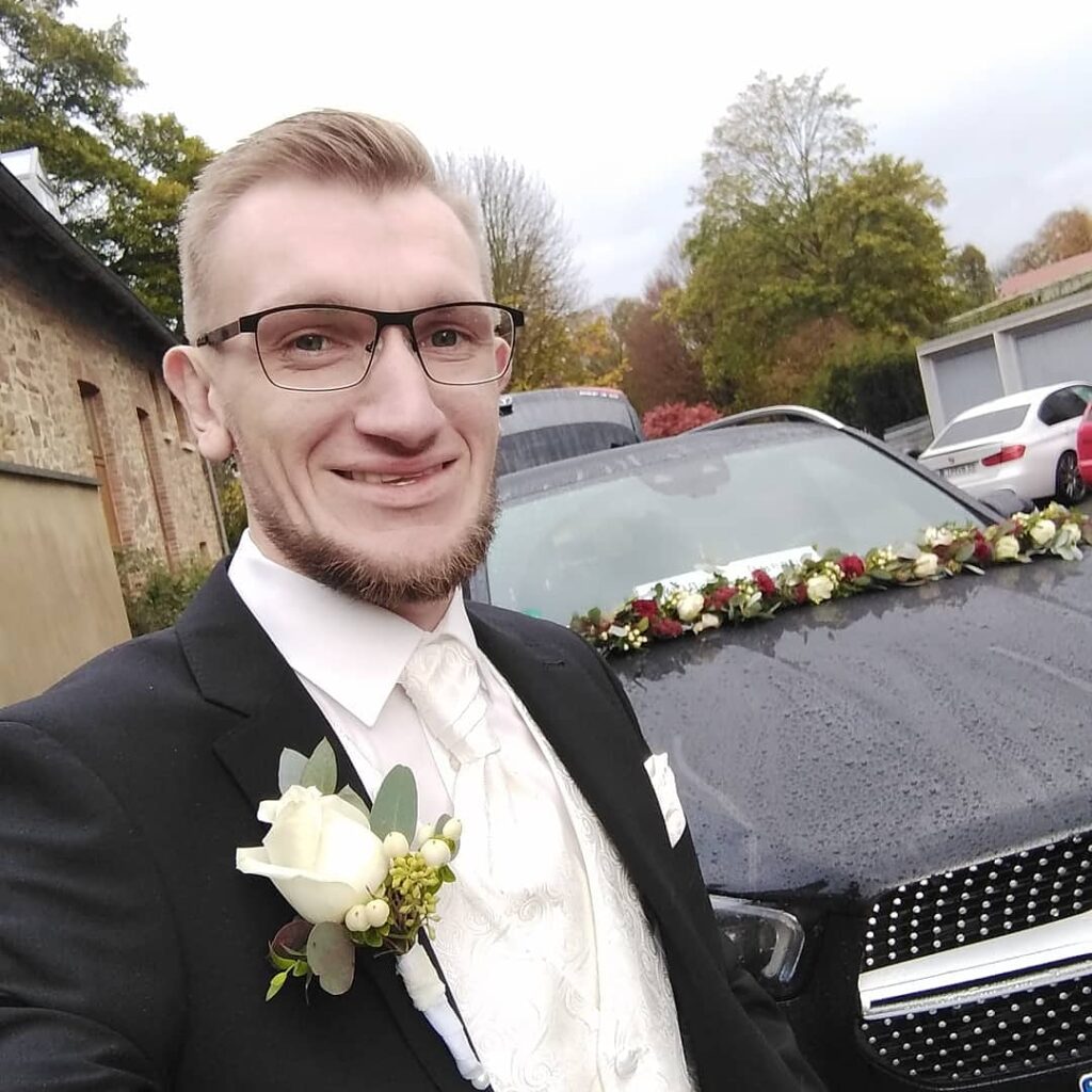 Ein Mann in einem schwarzen Anzug und mit einer Blume am Revers lächelt in die Kamera. Neben ihm steht ein schwarzes Auto mit Blumen auf der Motorhaube.