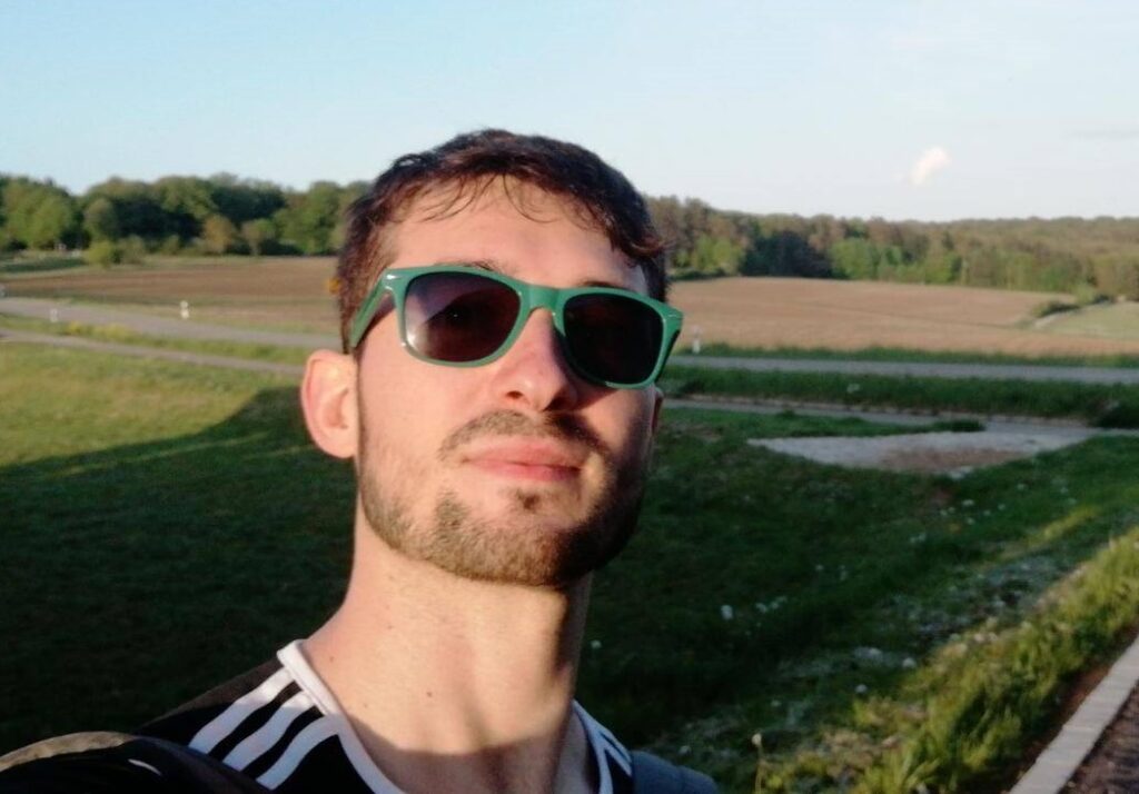 Ein Mann mit kurzen Haaren und grünen Sonnenbrille steht vor einer grünen Wiese und einem Feld.