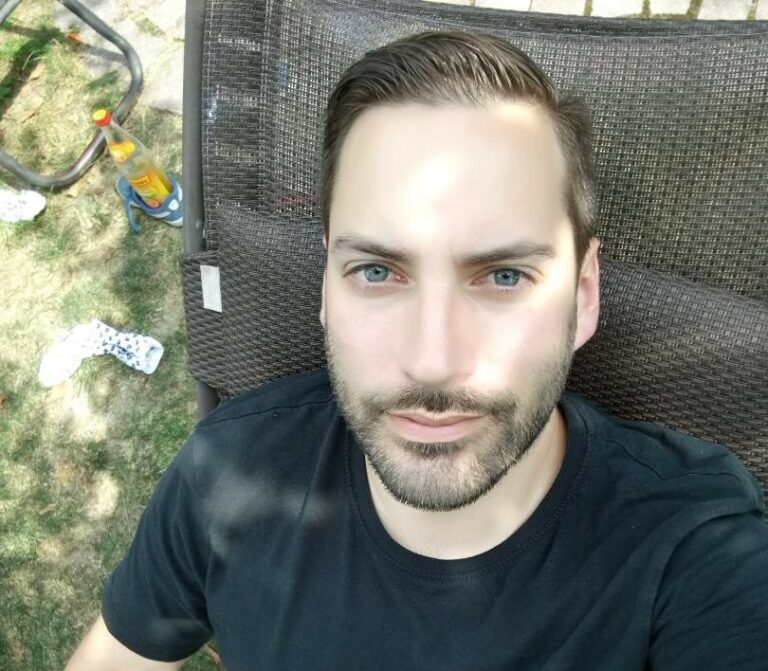 Ein Mann mit blauen Augen und einem gepflegten Bart sitzt auf einer Outdoor-Möbel. Er trägt ein schwarzes T-Shirt.