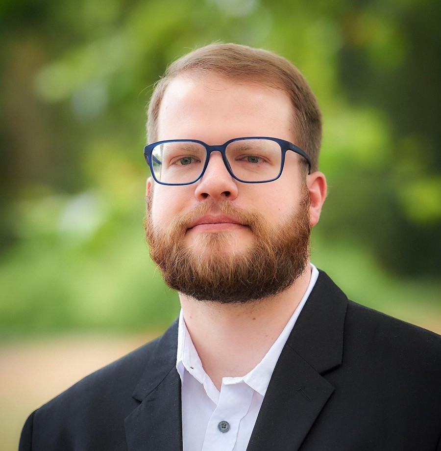 Ein Mann mit Brille und Bart, der im Freien steht. Er trägt ein weißes Hemd und einen schwarzen Anzug.