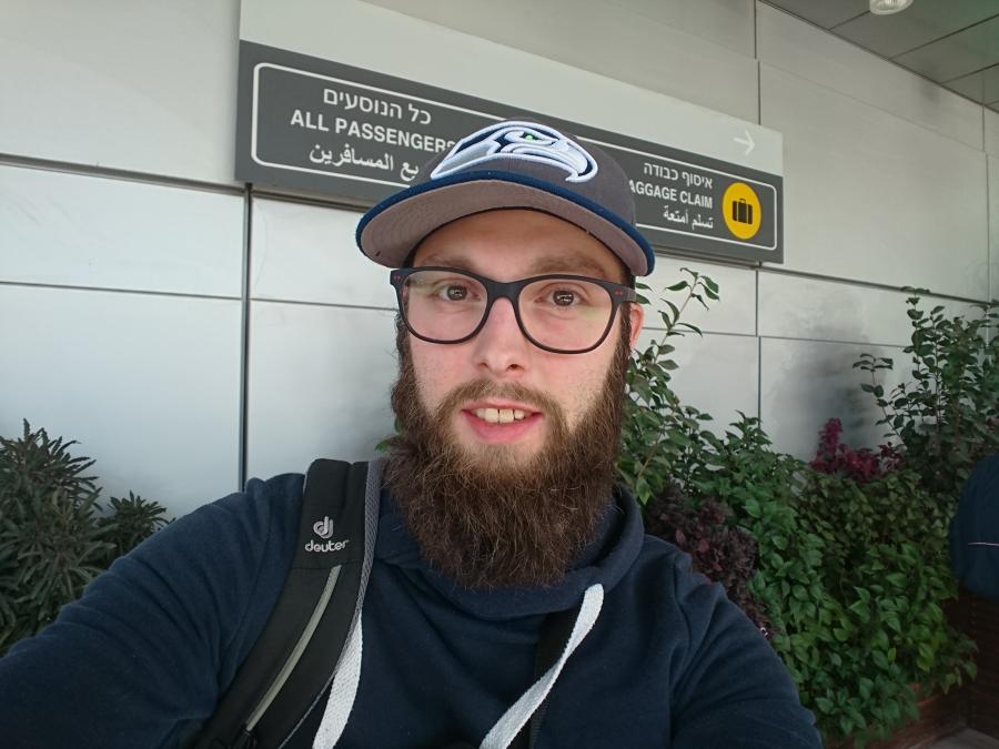 Ein Mann mit einem Bart und einer Kappe steht vor einem Flughafenschild.