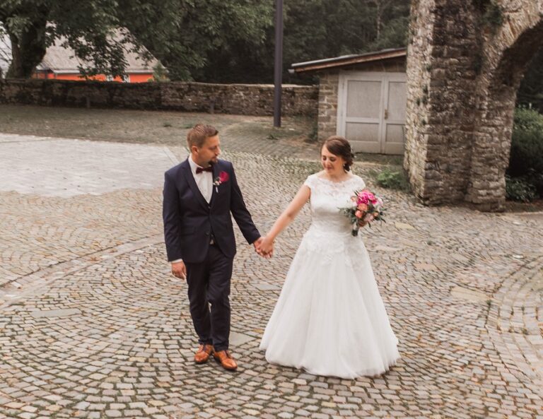 Ein frisch vermähltes Paar geht händchenhaltend über eine Kopfsteinpflasterstraße. Die Braut trägt ein weißes Kleid und einen Blumenstrauß.