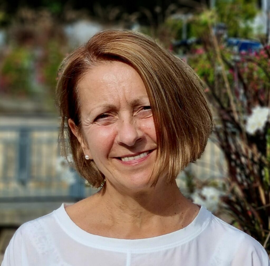 Eine lächelnde Frau im Garten, kurzhaarig und weiß gekleidet. Sie steht vor einer blühenden Pflanze.