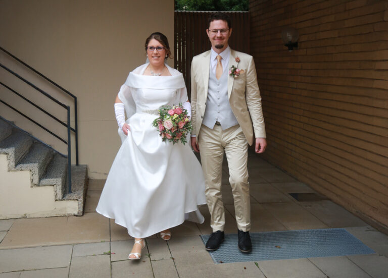 Ein frisch verheiratetes Paar, die Braut in weißem Kleid mit Blumenbouquet, der Bräutigam in beige-farbenem Anzug.