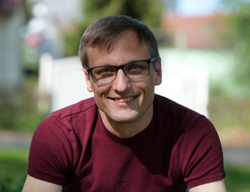 Ein Mann mit kurzen Haaren und Brille trägt ein rotes T-Shirt. Er lächelt freundlich.