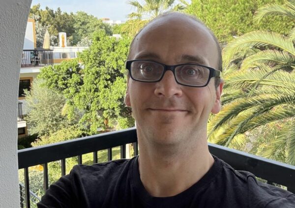 Ein Mann mit Brille steht auf einem Balkon mit Blick auf Bäume und ein Gebäude. Er trägt ein schwarzes T-Shirt.